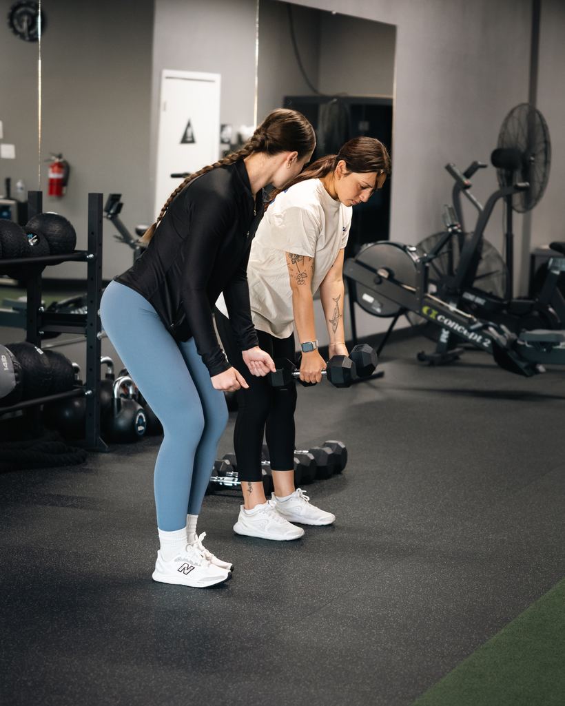 A coach demonstrating a dumbbell hinge alongside a client during a one-on-one evaluation at Merced Elite Gym