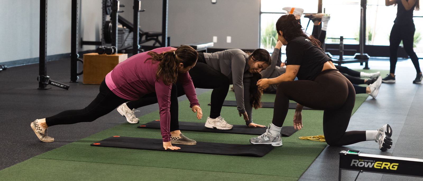 A personal trainer kneeling to correct a client's stretch position during a women's semi-private strength session at Merced Elite Gym in Merced, CA