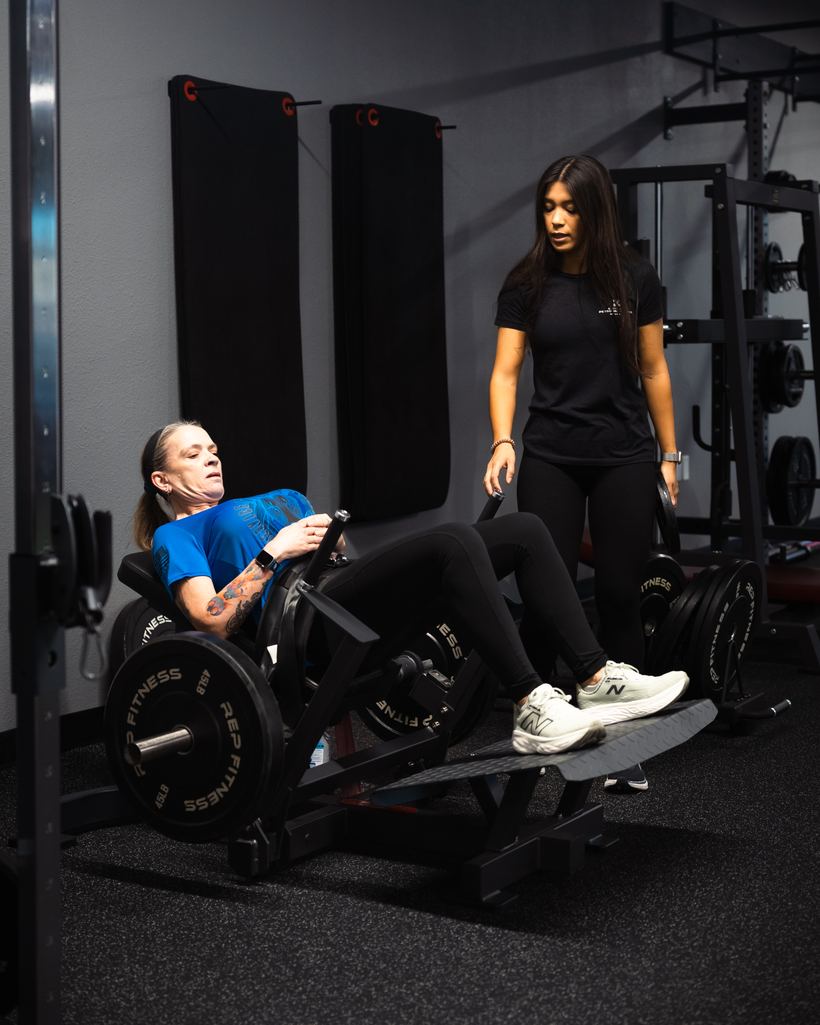 A woman in her fifties training on the leg press with her coach beside her at Merced Elite Gym in Merced, CA