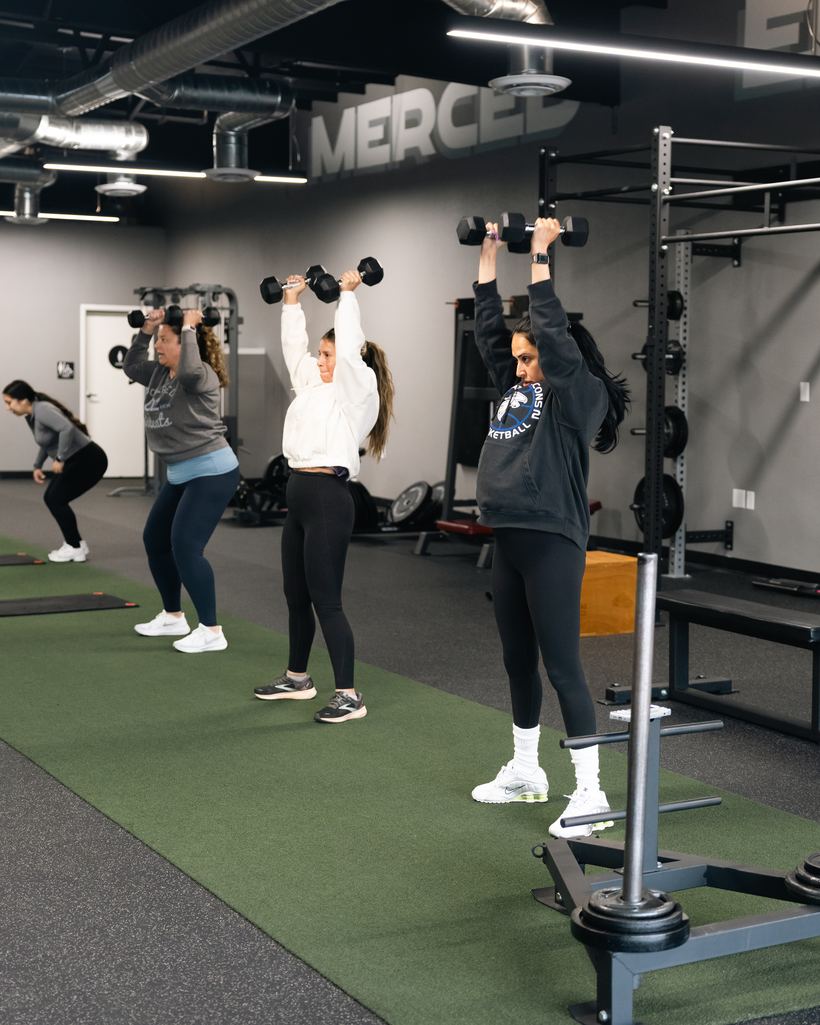 Four women pressing dumbbells overhead at different weights during a semi-private strength session at Merced Elite Gym